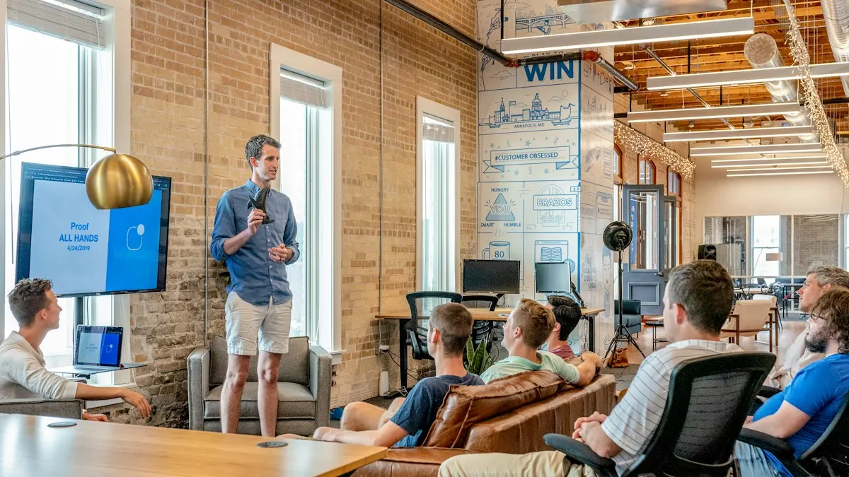A man stands and speaks to a group seated on sofas and office chairs in a brick-walled open office with a wall screen
