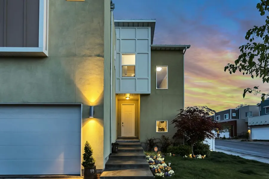 Two-story green stucco home with an arched garage, a lit entry and a colorful sky at dusk