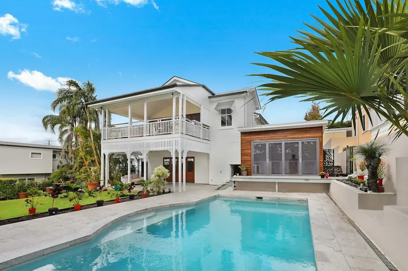 White two-story house with a wraparound upper veranda, a timber-clad extension, tropical plants and a pool in the foreground