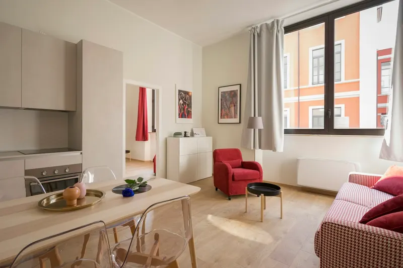 Bright open-plan apartment with a pale kitchen, a dining table with clear chairs, a red armchair and a large window onto a neighboring building