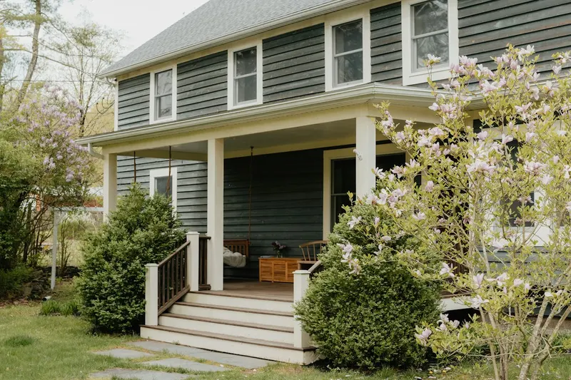 Two-story home with gray-green clapboard siding, a deep covered front porch with a swing, white steps and a flowering magnolia