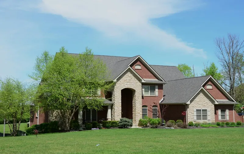 Large two-story brick home with a tall arched stone entry, gabled roofs, young trees and a wide green lawn