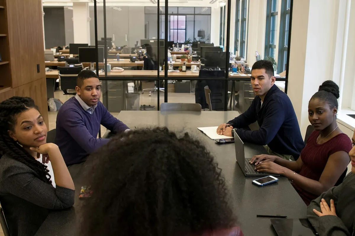 A group of young adults sit around a dark meeting table in an open office, one typing on a laptop while the others listen