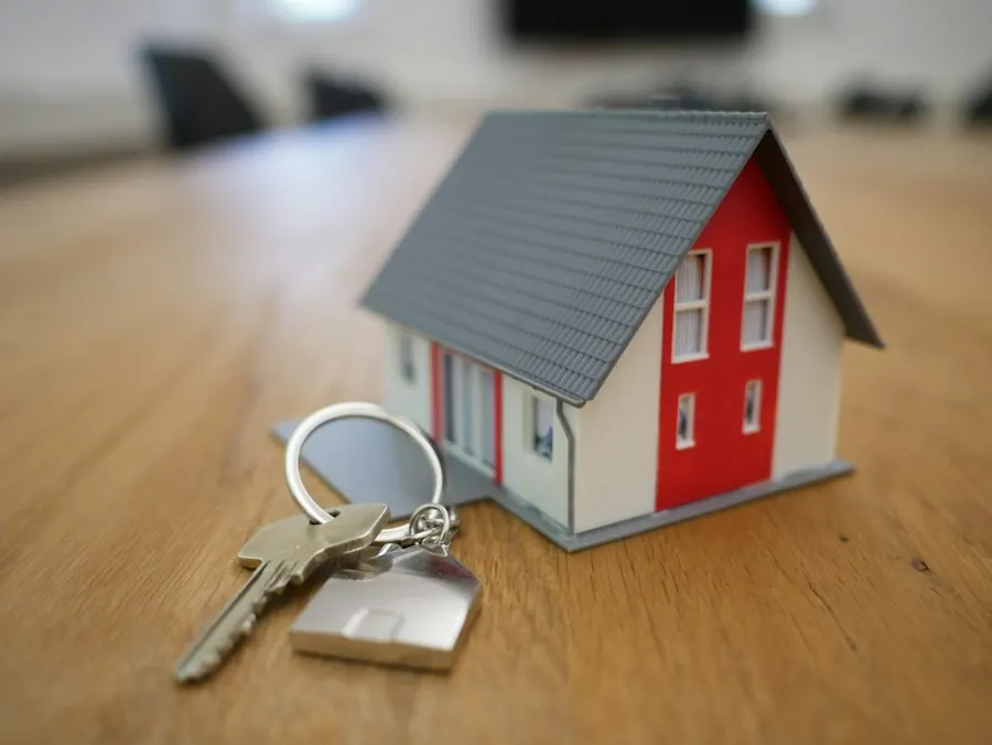 A small model house with a red front and a set of house keys on a wooden table