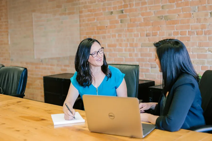 Two women talk across a wooden table with a laptop and a notepad in a brick-walled room
